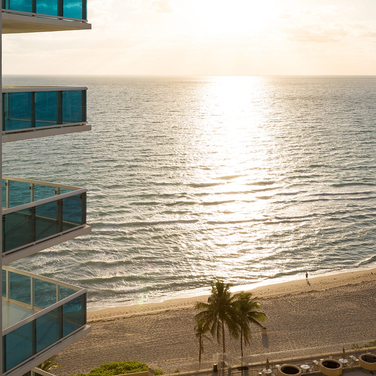 Sunset over the sea, seen past the glass balconies of a beachfront apartment block with palm trees below.